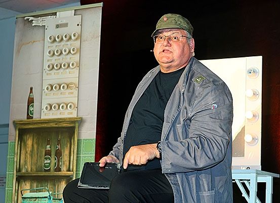 Ein älterer Mann sitzt auf einer Bühne und hält ein Tablet. Hinter ihm steht ein Holzregal mit Flaschen und eine Lampe auf der rechten Seite.