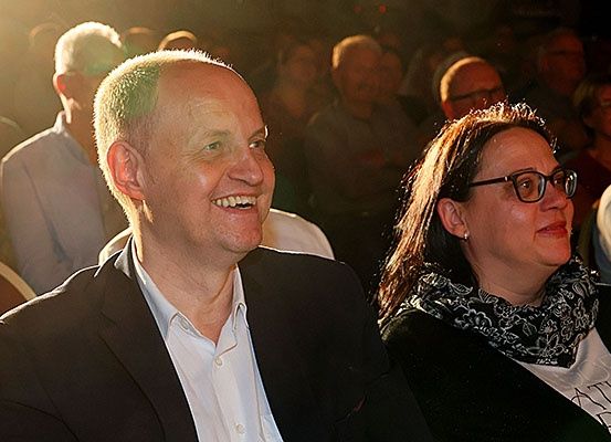 Ein Mann und eine Frau lächeln und schauen in die Kamera, sie sitzen im Publikum bei einer Veranstaltung.