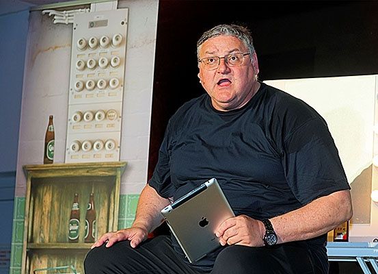 Ein Mann sitzt auf der Bühne, hält ein iPad und hat Regale mit Flaschen und ein Schalttafel mit Knöpfen hinter sich.