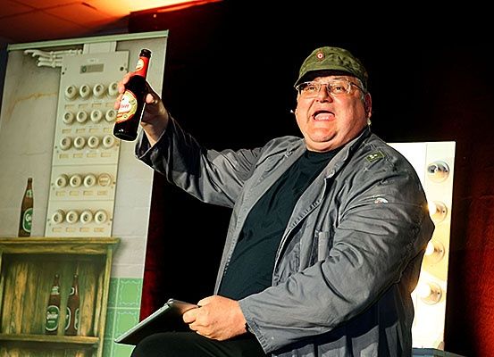 Ein Mann in einer grünen Mütze und Brille sitzt auf einem Stuhl und hält eine Bierflasche in seiner erhobenen rechten Hand. Er scheint auf einer Bühne zu sein. Hinter ihm befindet sich eine bemalte Kulisse einer Wand mit Regalen und einer Reihe von Flaschen.