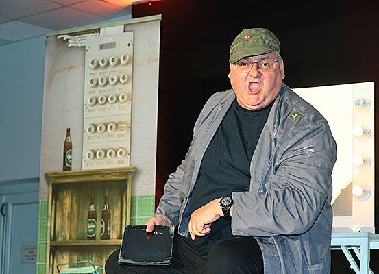 Ein Mann sitzt auf einem Stuhl und hält ein Tablet in der Hand, während er es ansieht und eine überraschte Miene macht. Er trägt einen grünen Hut und eine Brille. Hinter ihm steht ein Holzregal mit Flaschen darauf und eine weiße Box mit runden Knöpfen.