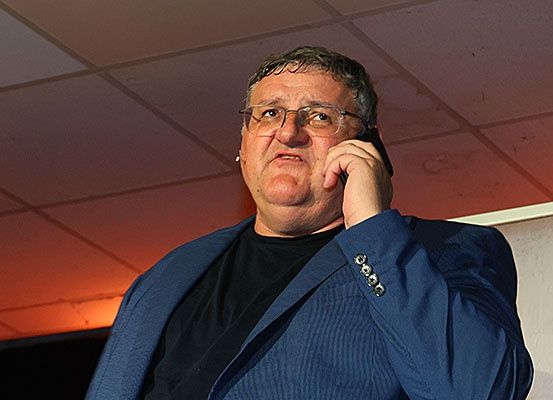 Ein Mann in einem blauen Blazer spricht am Handy und steht gegen eine weiße Wand mit rot beleuchteter Decke.
