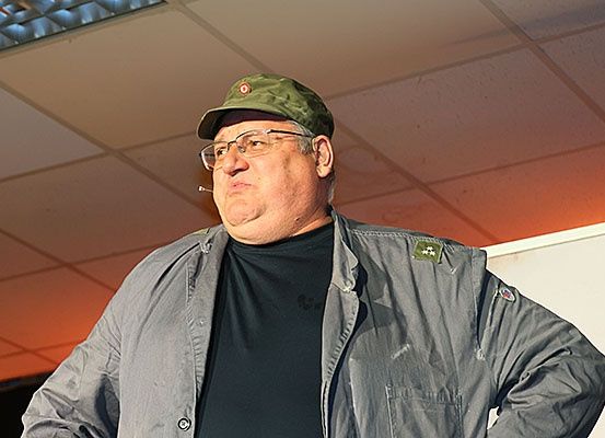 Ein älterer Mann mit grüner Mütze und Brille steht vor einer weißen Wand. Er hat eine Zigarette im Mund und ist in einem schwarzen Hemd und einer grauen Jacke gekleidet.