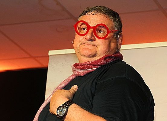 Ein Mann in einem schwarzen Shirt, roten Brille und einem Schal steht vor einem Whiteboard und schaut zur Kamera.