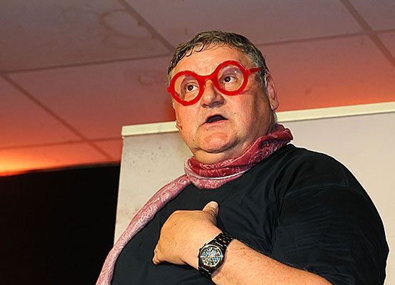 Ein älterer Mann mit roter Brille und Schal steht und scheint zu sprechen, während er seine Brust berührt.