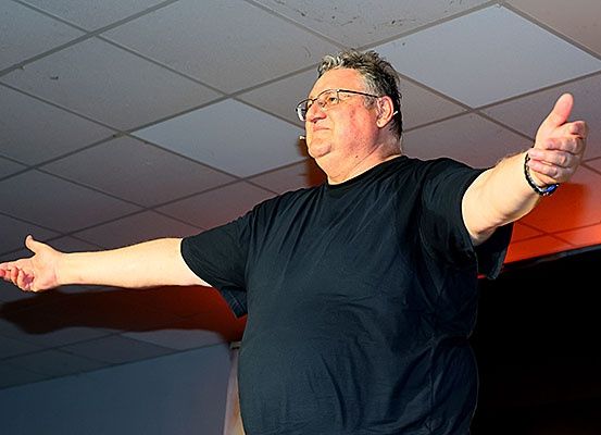 Ein Mann mit Brille und schwarzem T-Shirt steht auf der Bühne, gestikuliert mit beiden Händen und hält möglicherweise eine Rede oder Präsentation. Der Hintergrund ist schwach beleuchtet.