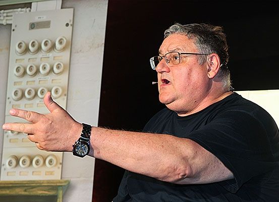 Ein Mann mit grauem Haar und Brille, der ein schwarzes T-Shirt und eine Uhr trägt, gestikuliert mit der Hand und raucht eine Zigarette.