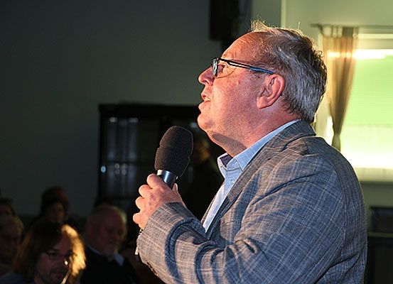 Ein Mann mit Brille und grauem Anzug spricht in ein Mikrofon in einem Raum mit zuschauenden Leuten.