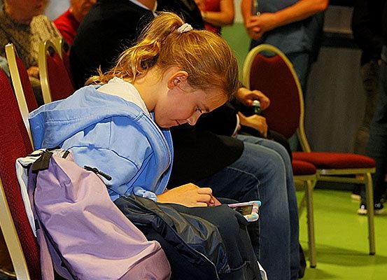 Ein junges Mädchen sitzt auf einem Stuhl und schaut auf ihr Handy, während andere in der Nähe sind.