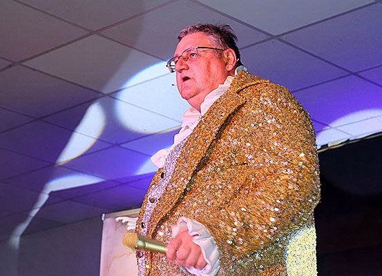 Ein Mann in einem goldenen Pailletten-Anzug steht auf der Bühne, hält ein Mikrofon und tritt unter einem Scheinwerfer auf.