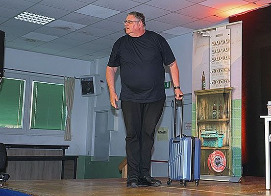 Ein Mann steht mit einem Koffer auf der Bühne, trägt eine Brille und ein schwarzes T-Shirt. Hinter ihm befindet sich eine Wand mit einem Holzregal, das verschiedene Gegenstände präsentiert.