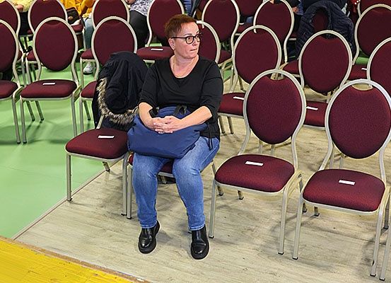 Eine Frau mit Brille und schwarzem Shirt sitzt in einem Stuhl mit verschränkten Händen auf dem Schoß. Sie hat eine blaue Tasche auf dem Schoß. Die Stühle sind dunkelrot mit weißen Gestellen.