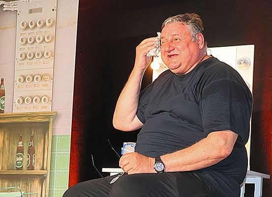 Ein älterer Mann in einem schwarzen T-Shirt sitzt auf einem Stuhl, hält ein Taschentuch in der Hand und scheint zu weinen. Er trägt eine Uhr am linken Handgelenk. Hinter ihm ist ein Regal mit zwei Flaschen zu sehen, und ein Spiegel reflektiert eine Lampe.