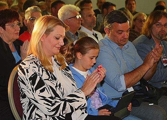 Eine Gruppe von Menschen sitzt in einem Saal. Eine Frau und ein Mädchen klatschen in die Hände. Ein Mann hält einen Laptop.