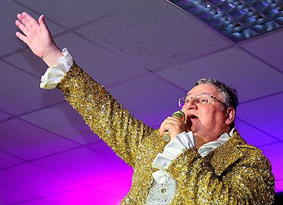 Ein erwachsener Mann in einem goldglänzenden Paillettenanzug und mit Brille singt in ein Mikrofon auf einer Bühne mit lila Beleuchtung.