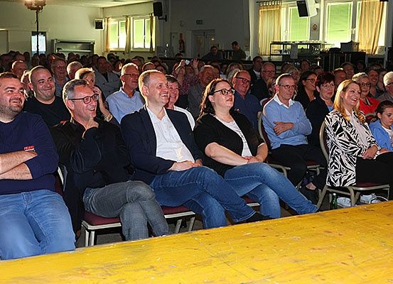 Eine Gruppe von Menschen sitzt in Stühlen in einem Raum, lächelt und schaut zur Kamera. Die Stühle sind in Reihen angeordnet, und der Raum hat gelb gestrichene Wände und Fenster.