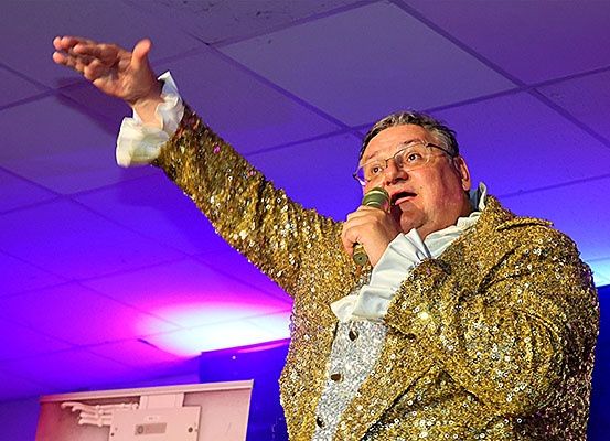Ein Mann in einem goldenen Pailletten-Anzug mit weißem Innenshirt und Brille singt in ein Mikrofon, während er die rechte Hand hebt.