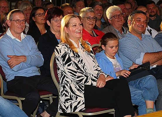Eine Gruppe von Erwachsenen und Kindern sitzt in einem Raum, möglicherweise bei einer Versammlung. Eine Frau in einer Zebra-Jacke lächelt.