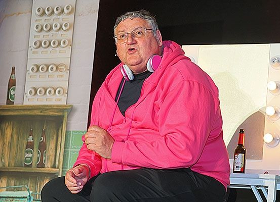 Ein älterer Mann sitzt auf einem Stuhl und trägt eine pinke Kapuzenjacke und schwarze Hose. Er trägt eine Brille und Kopfhörer. Eine Flasche steht auf dem Tisch neben ihm. Hinter ihm ist ein Regal mit Flaschen und eine Wand mit einem Bild.
