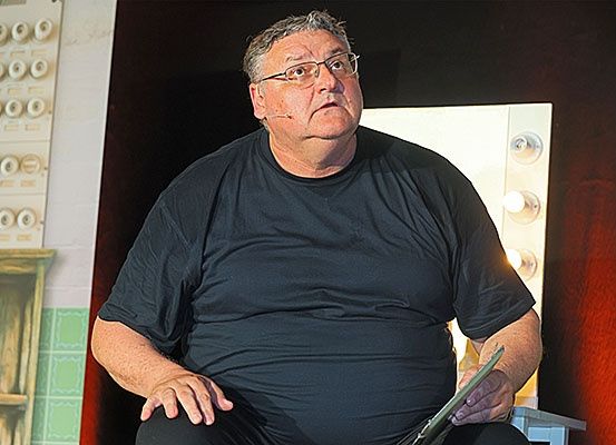 Ein älterer Mann mit Brille und schwarzem T-Shirt sitzt auf einem Hocker und hält ein Tablet in seinem Schoß. Er sieht nachdenklich aus, mit einem Mikrofon im Ohr. Hinter ihm ist eine rote Wand mit einer weißen Leuchte.
