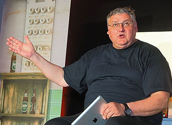 Ein Mann mit Brille und schwarzem T-Shirt spricht zu einem Publikum, während er auf einem Stuhl sitzt und ein iPad hält. Hinter ihm steht ein Holzregal mit Flaschen und eine dekorative Wand.