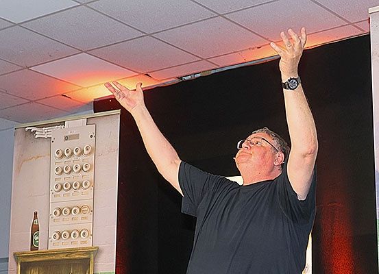 Ein Mann steht mit erhobenen Armen auf einer Bühne, trägt ein schwarzes T-Shirt, eine Brille und eine Uhr. Hinter ihm befindet sich ein schwarzer Hintergrund mit einer roten Leuchte. Links steht ein Holzregal mit einer Flasche darauf.