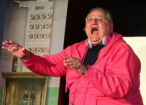 Ein älterer Mann in einer rosa Kapuzenjacke und mit Brille spricht in ein Mikrofon, mit offenem Mund und erhobenen Händen. Hinter ihm befindet sich eine weiße Wand mit einer Schalttafel und einem Holzrahmen mit Bierflaschen.