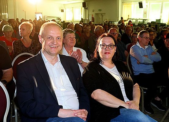 Eine Gruppe von Menschen sitzt in einem Saal, einige lächeln und klatschen. Ein Mann und eine Frau sitzen in der ersten Reihe, mit vielen anderen dahinter. Der Raum ist hell mit Sonnenlicht.