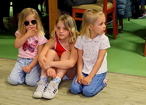 Drei junge Mädchen sitzen auf einem Holzboden, wahrscheinlich in einem Studio. Sie tragen Freizeitkleidung. Ein Mädchen trägt eine Sonnenbrille.