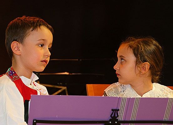 Zwei junge Kinder, ein Junge und ein Mädchen, stehen sich gegenüber und scheinen in ein Gespräch oder eine Aufführung vertieft zu sein. Der Junge trägt ein traditionelles Kostüm.
