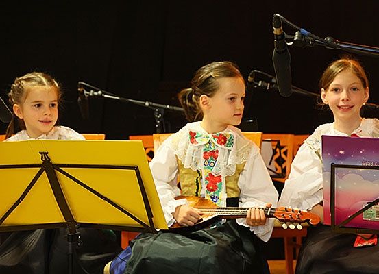 Drei Mädchen in traditioneller Kleidung spielen auf der Bühne Musikinstrumente. Ein Mädchen in der Mitte spielt eine Gitarre, während die anderen Musikblätter vor sich haben. Mikrofone sind hinter ihnen aufgestellt.