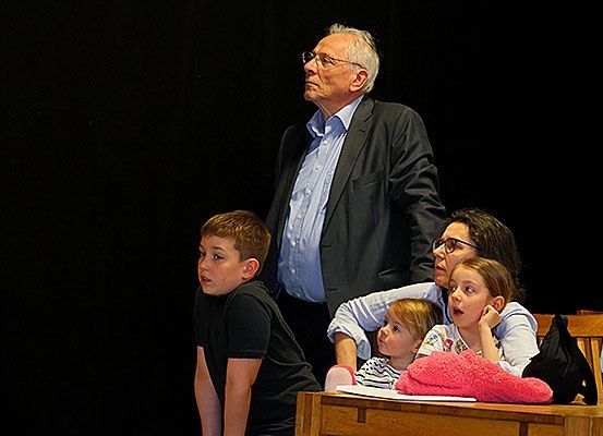 Ein älterer Mann in einem Anzug steht auf der Bühne und schaut nach oben. Eine vierköpfige Familie sitzt vor ihm an einem Tisch. Die Frau und die Kinder schauen in seine Richtung.