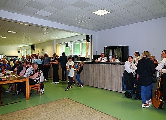Eine Gruppe von Menschen versammelt sich in einem großen Raum, einige stehen und andere sitzen an Tischen. Eine Theke mit Leuten dahinter und eine Person, die eine Gitarre hält.