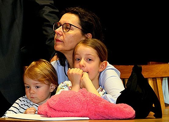 Eine Frau und zwei Kinder sitzen aufmerksam an einem Tisch, möglicherweise in einem Gerichtssaal. Die Frau trägt eine Brille, und die Kinder scheinen auf etwas vor ihnen konzentriert zu sein. Eine rosa Decke liegt auf dem Tisch.