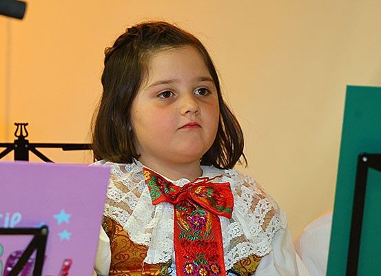 Ein junges Mädchen in einem traditionellen Kleid mit einer roten Schleife steht vor einem Notenständer, möglicherweise bei einer Aufführung. Hinter ihr befindet sich eine lila Karte mit Sternen und einem Mikrofon.