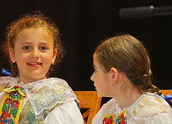 Zwei junge Mädchen in traditionellen Kostümen sitzen auf einer Holzbank. Das Mädchen auf der linken Seite lächelt, während das Mädchen auf der rechten Seite wegschaut. Sie haben beide geflochtene Haare mit blauen Bändern.