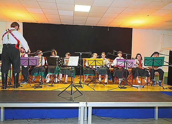 Eine Gruppe junger Musiker tritt mit ihren Instrumenten auf der Bühne auf. Sie sitzen mit Notenständern. Ein Mann steht vorne, möglicherweise leitet er sie.