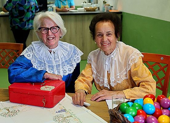Zwei ältere Frauen sitzen an einem Tisch, eine hält eine rote Box und lächelt. Bunte Eier in einem Korb auf dem Tisch. Dahinter steht eine Person in der Nähe einer Theke.