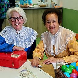 Zwei ältere Frauen sitzen an einem Tisch, eine hält eine rote Box und lächelt. Bunte Eier in einem Korb auf dem Tisch. Dahinter steht eine Person in der Nähe einer Theke.