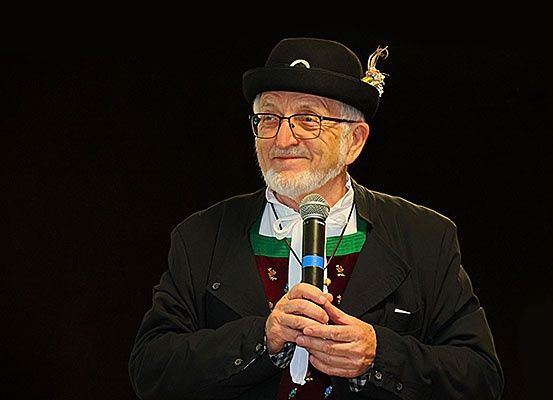 Ein älterer Mann in einem schwarzen Hut und einer Brille, der einen schwarzen Mantel und ein weißes Hemd trägt, hält ein Mikrofon. Er lächelt und hält wahrscheinlich eine Rede.