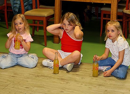 Drei junge Mädchen sitzen auf dem Boden, jede hält eine Flasche Orangenlimonade. Sie scheinen sich in einem Spielzimmer zu befinden, mit Stühlen und einem Holztisch hinter ihnen.