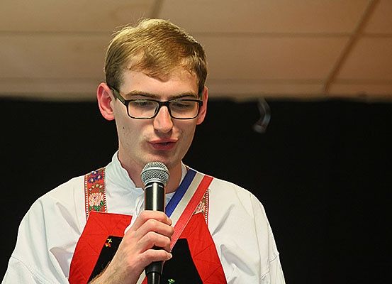 Ein junger Mann mit Brille und einer Medaille um den Hals hält ein Mikrofon, möglicherweise bei einer Bühnenaufführung.