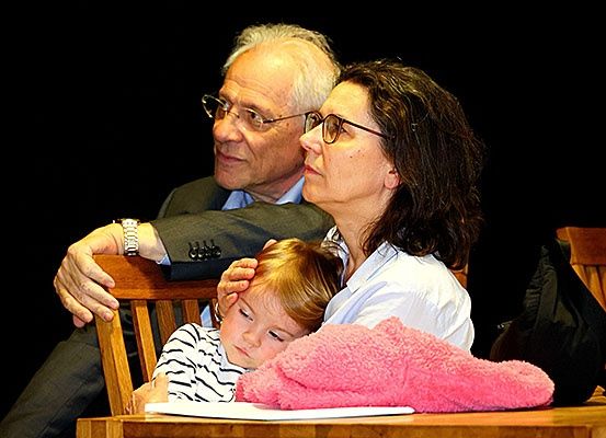 Ein älterer Mann mit Brille tröstet ein schlafendes Kind, während eine Frau in der Nähe sitzt. Das Kind ruht auf dem Schoß der Frau.