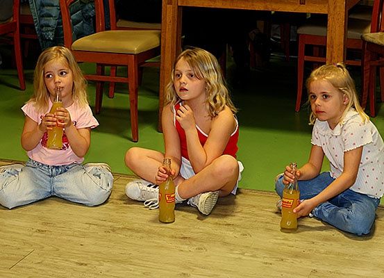 Drei kleine Mädchen sitzen auf dem Boden, jedes hält eine Flasche Limonade. Sie sind von Holzstühlen und einem Tisch umgeben. Der Boden ist grün und der Raum ist schwach beleuchtet.