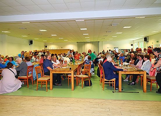 Eine große Gruppe von Menschen sitzt an Tischen in einem Saal, viele sind älter, und einige tragen Hüte. Flaschen und Becher stehen auf den Tischen. Der Raum hat weiße Wände und einen grünen Boden.