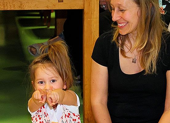 Ein kleines Mädchen mit blonden Haaren zeigt auf etwas und lächelt, während eine Frau in einem schwarzen T-Shirt sie ansieht.