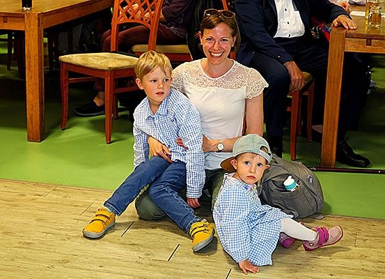 Eine Frau und zwei Kinder sitzen auf einem Holzboden und lächeln. Die Frau hält einen Jungen auf dem Schoß. Ein anderer Junge sitzt neben ihr. Hinter ihnen sitzt ein Mann im Anzug an einem Tisch.