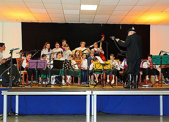 Eine Gruppe von Kindern und Erwachsenen spielt auf der Bühne mit Musikinstrumenten. Die Bühne ist mit einem blauen Hintergrund und einem schwarzen Vorhang dekoriert.