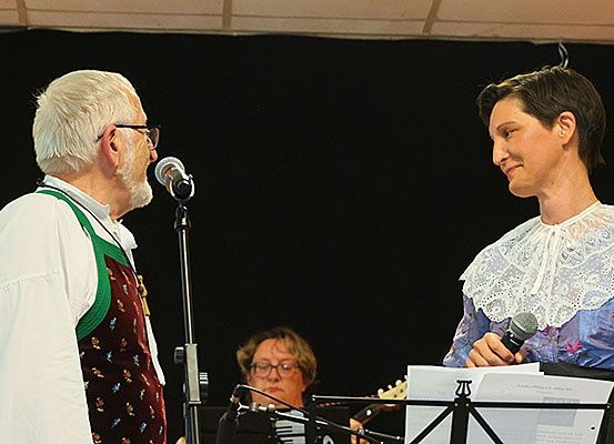 Ein Mann und eine Frau, beide mit Mikrofonen in der Hand, scheinen auf der Bühne zu stehen. Hinter ihnen spielt ein Musiker Akkordeon. Ein Notenständer mit Noten steht vor der Frau.