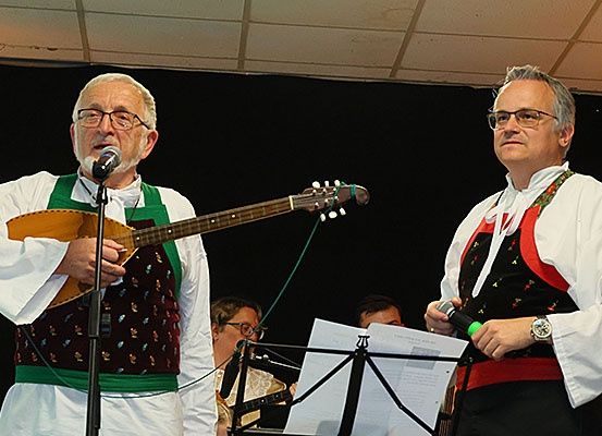 Zwei Männer stehen auf der Bühne, einer mit einer Gitarre, der andere mit einem Mikrofon, in traditionellen Volkstrachten, begleitet von Musikern im Hintergrund.